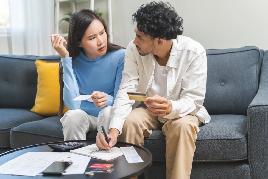 A stressed couple sits on the couch with credit cards and unpaid bills on the table in front of them