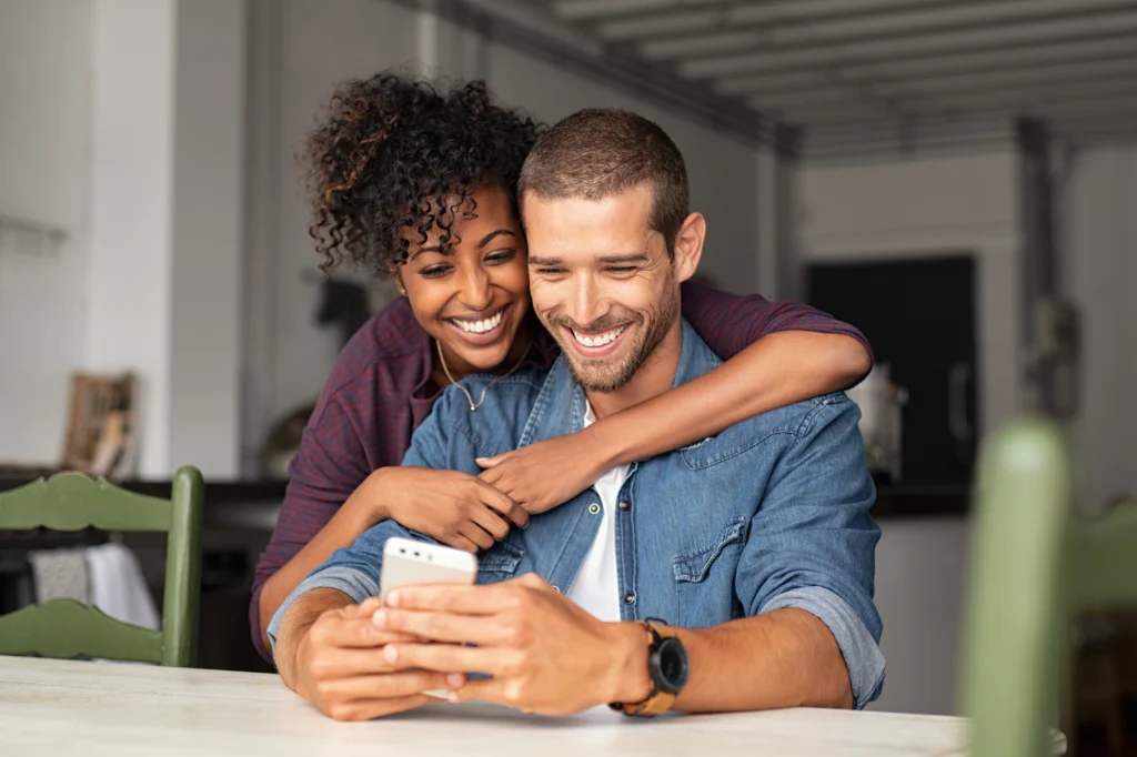 A man smiles at his phone while a woman with an arm around him looks over his shoulder