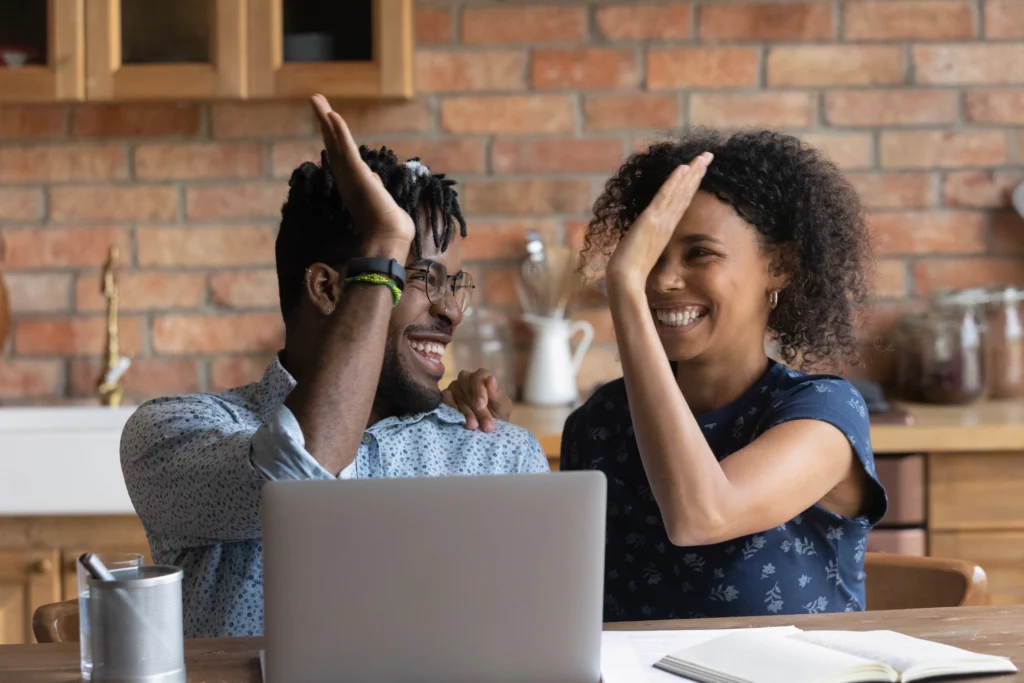 Smiling couple sitting at a table in front of a laptop high five after paying off their debt