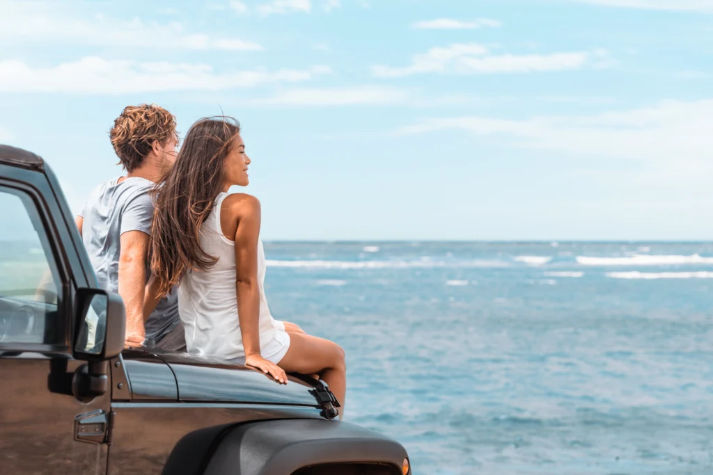 Smiling couple sits on the bonnet of their car looking out at the ocean