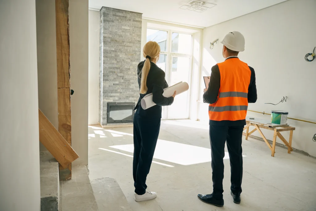 An architect in a hard hat and hi-vis vest stands in the living room of a recently build home, talking his client through it