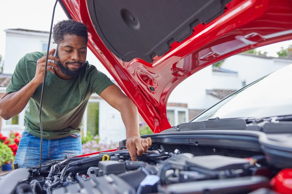 A man leans into the open hood of his car while on the phone to a mechanic
