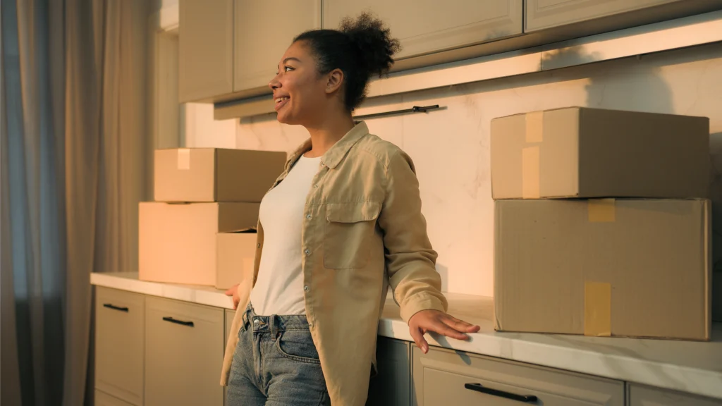 A happy woman stands in her kitchen surrounded by boxes after recently deciding whether to build a house vs buy an existing house