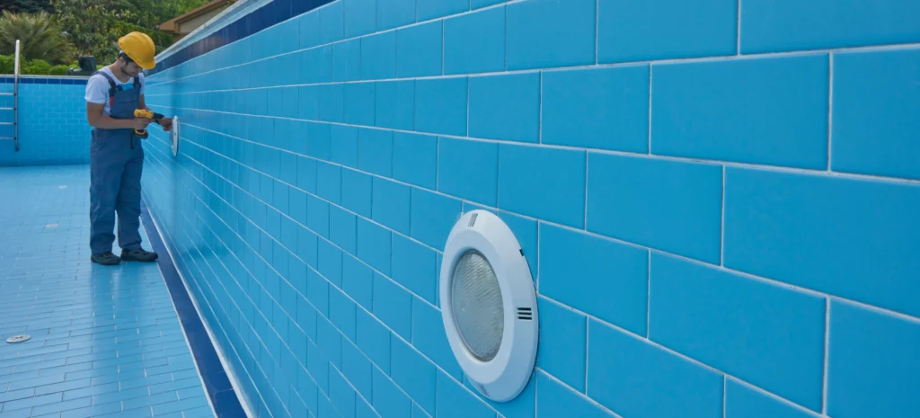A builder in overalls drills a light into the tiled wall of am empty pool
