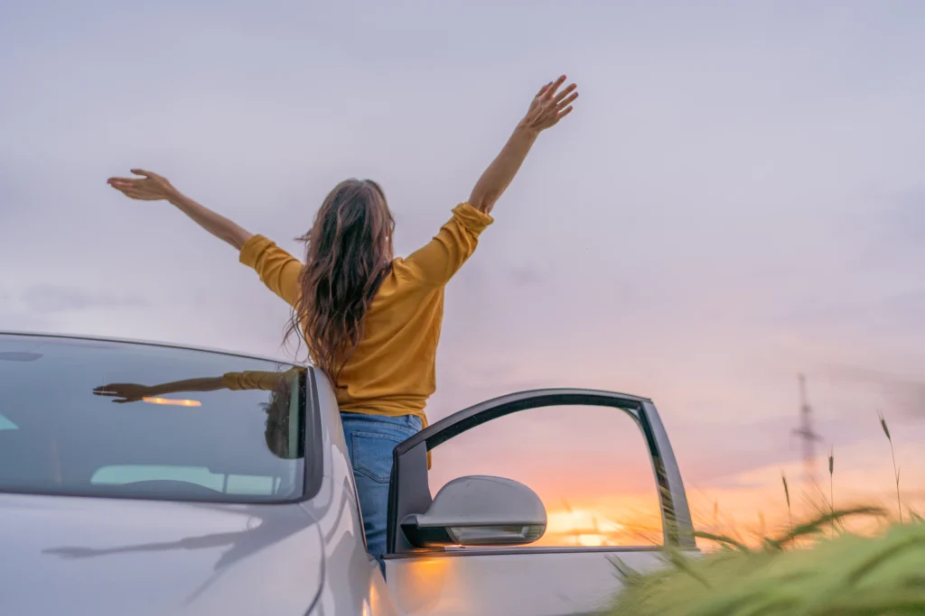 A woman leans out the drivers side of her car facing the sunset with her hands in the air