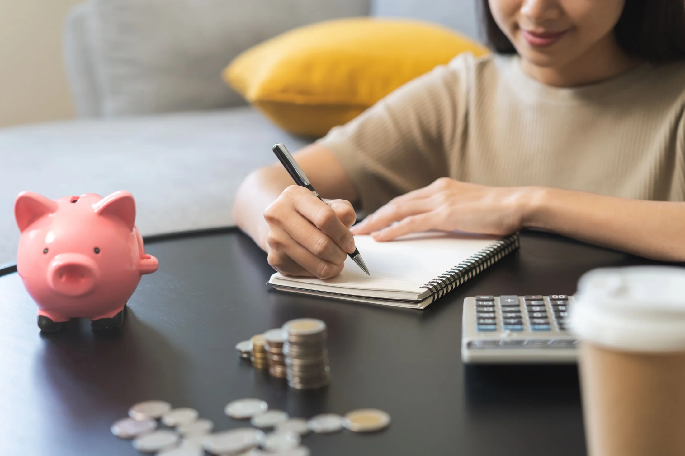 A woman sitting at a table writing her Christmas budget list with a pink piggy bank beside her, representing planning and saving for Christmas expenses.