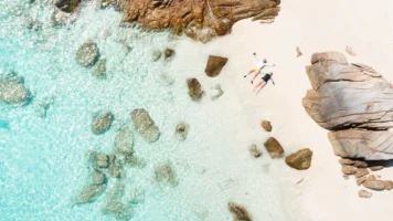 An aerial view of two people relaxing on a white-sand beach beside clear turquoise water and large coastal rocks.